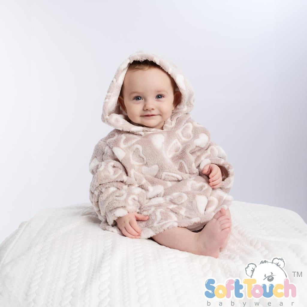 A baby wearing a beige hoodie with a heart pattern and hood, sitting on a similar colored soft surface.