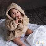 A baby wearing a beige hooded waffle texture jacket with a pocket, sitting on a blanket.