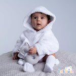 A baby wearing a white waffle hoodie with a hood pocket, paired with white pants, sitting and holding a gray toy.